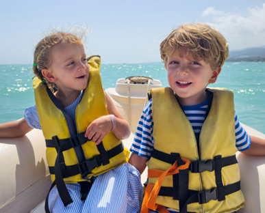 Two Young Kids In Lifejackets On The Back Or An Inflatable Boat With Dark Green Mountain Backdrop