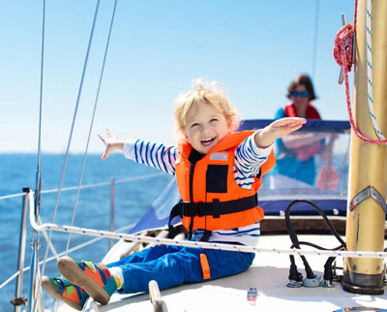 Boy In Life Jacket With Arms Outspread Sitting To The Side Of The Front Of A Yacht