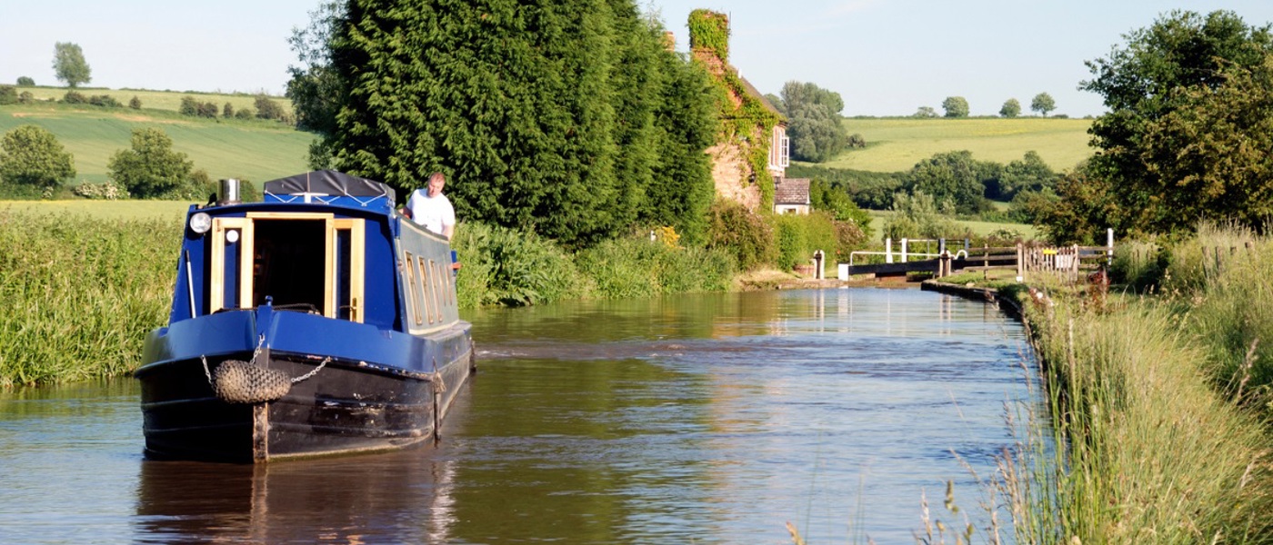 Man standing on a blue narrowboat on a canal in the sunshine